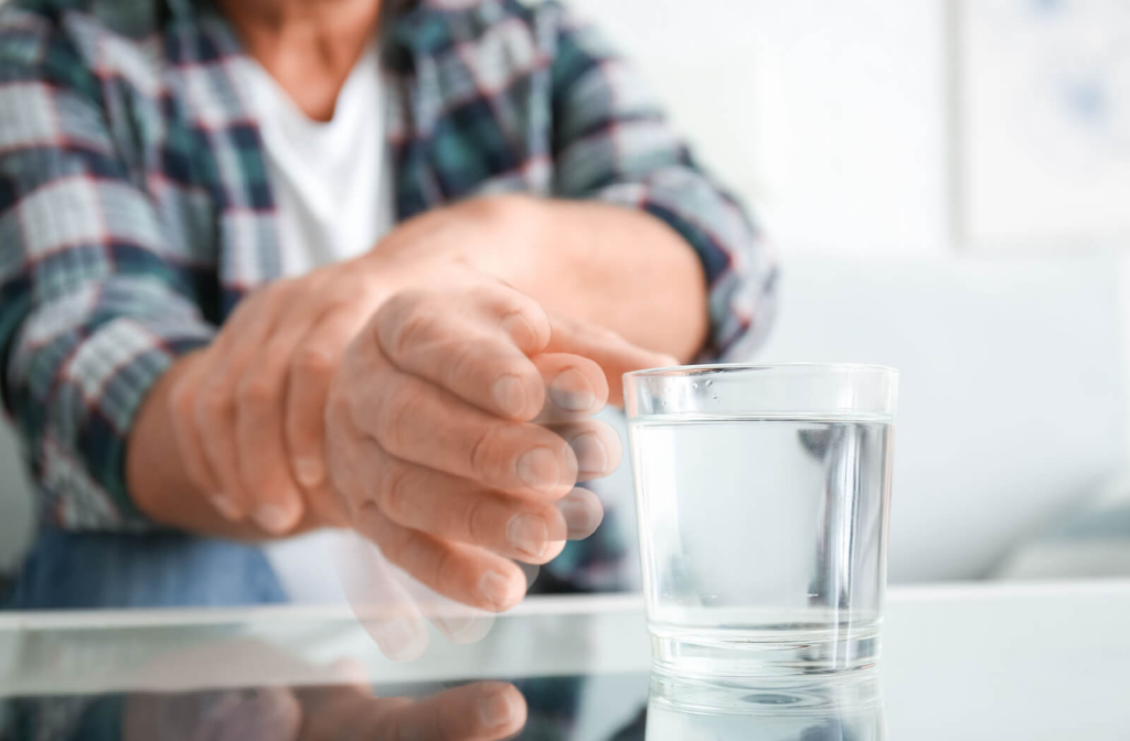 A senior man holds his wrist with his left hand to prevent tremors in his right hand while he reaches out for a glass of water.