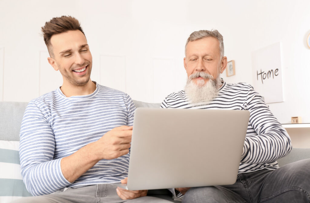 A senior man and a younger man sitting on a couch together looking at a laptop. Both are wearing striped shirts.