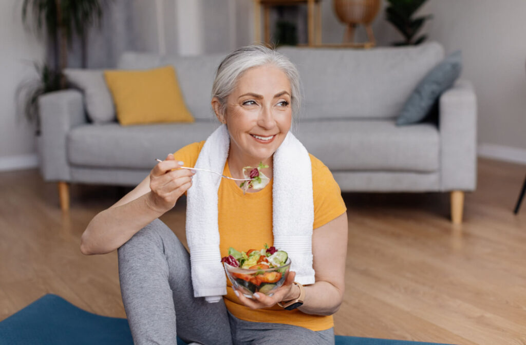senior woman enjoying a bowl of salad while seated on an exercise mat.