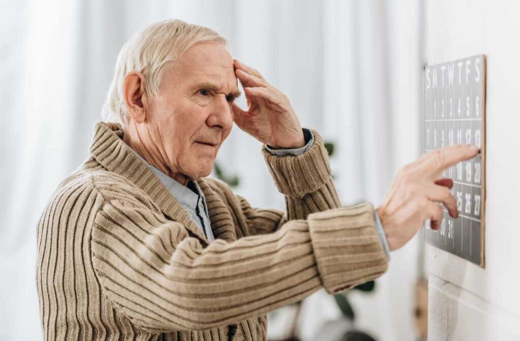 A senior man with dementia gently touches his forehead in an attempt to recall the date as he looks at a wall calendar