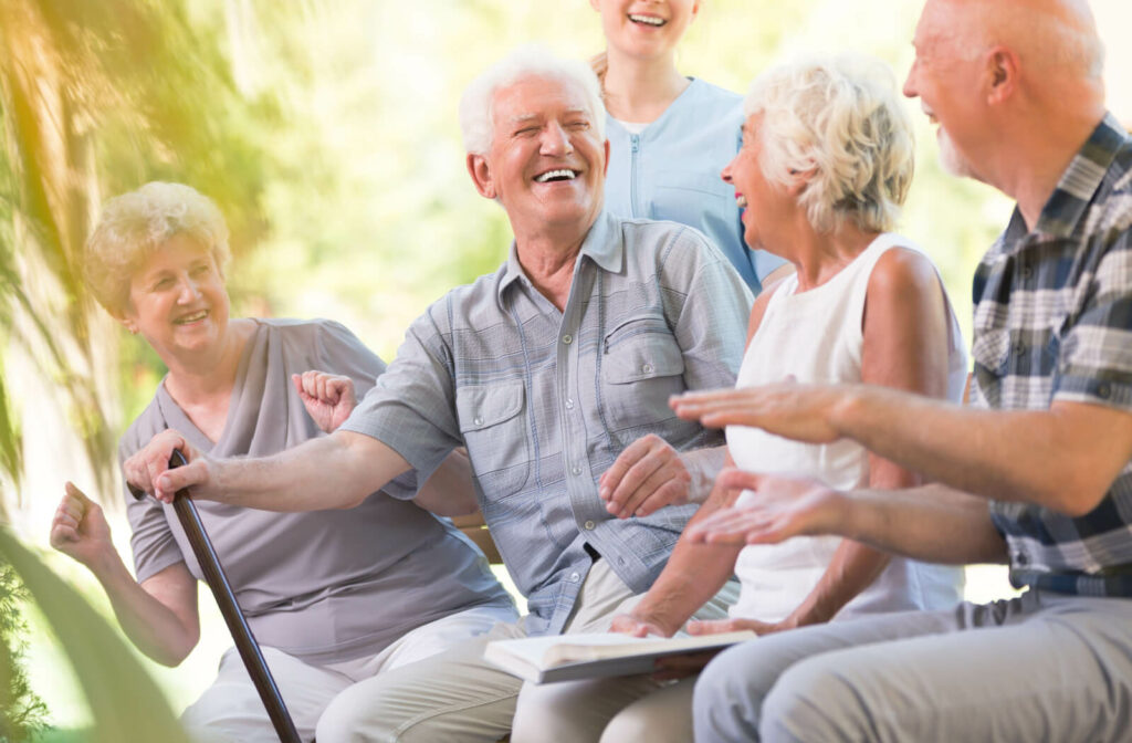 An older adult man with a cane smiles and laughs while sitting outside with a nurse and other older adults.
