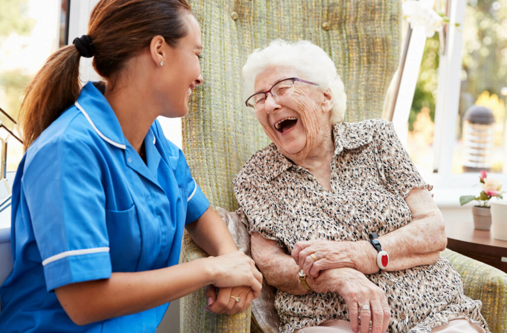 An older adult woman in a senior living facility sitting on a chair smiling and having a conversation with a nurse.